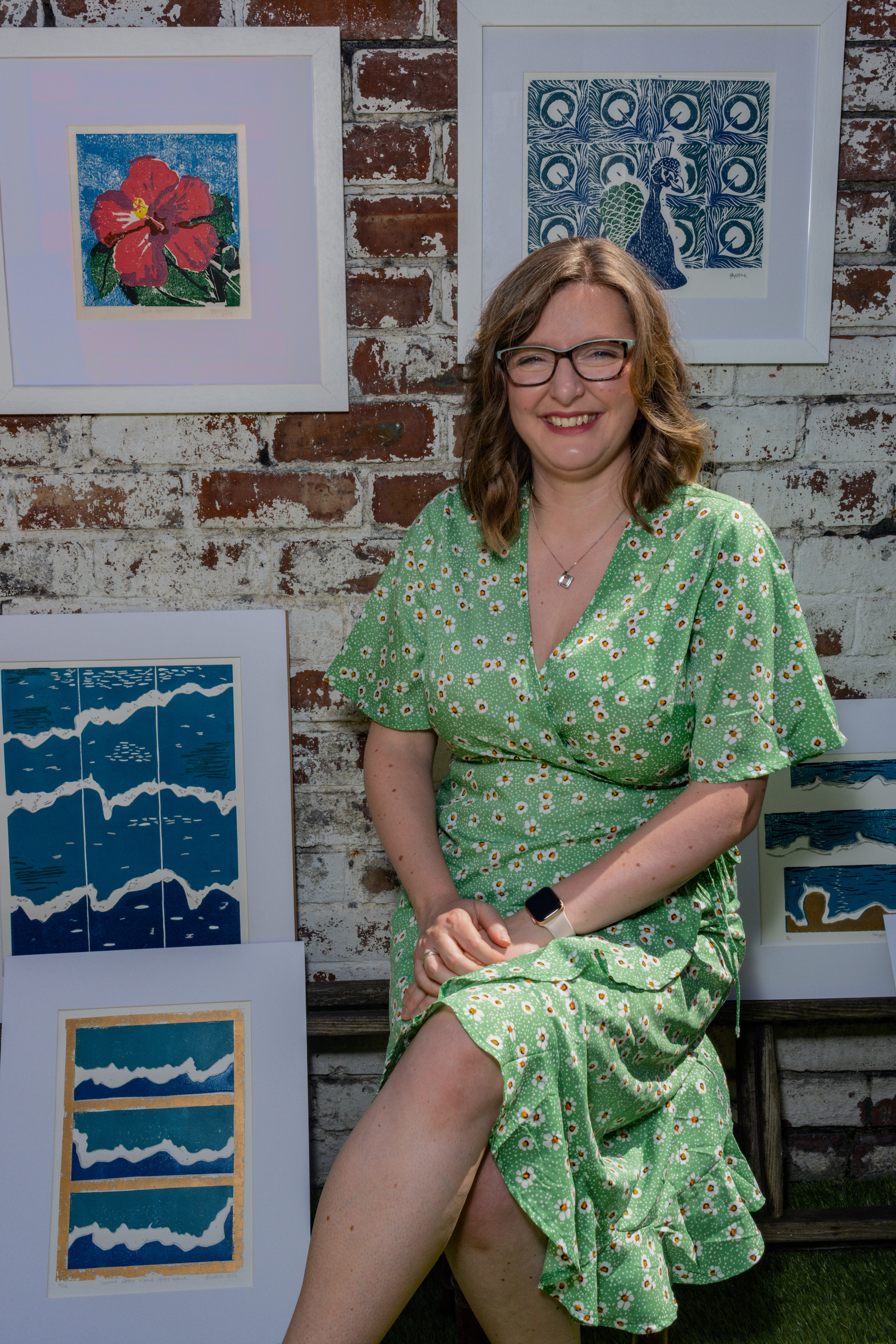 Heather Moore Newcastle Printmaker in a green floral dress in front of a brick wall covered in her framed artwork