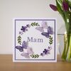 A square card stood next to a vase on a shelf. The white card has a purple background colour with a white foreground. The design features 4 cut out butterflies, 4 green leaf branches and 6 flowers placed in a repeating circular pattern.