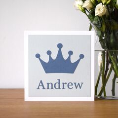 A photograph of a square card on a shelf next to a vase of flowers. The card is white with a light blue background and a darker blue crown shape on the front and a name underneath the crown.