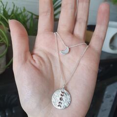 2 piece layered necklace on silver chain. circular silver coloured pendant with vertical moon phase cut out on the longer chain and a dainty shiny crescent moon on a short choker style chain