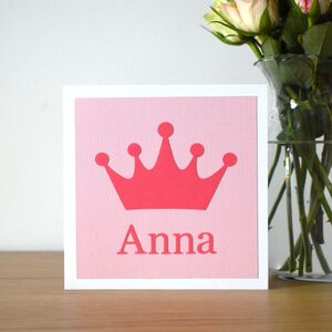 A photograph of a square card on a shelf next to a vase of flowers. The card is white with a light pink background and a darker pink crown shape on the front and a name underneath the crown.